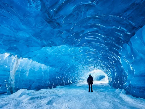 Inside a glowing blue glacier ice cave, with one person in a helmet seen from behind