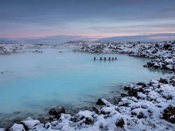 Steaming milky-blue geothermal lagoon among snowy lava rocks, with a few bathers far away