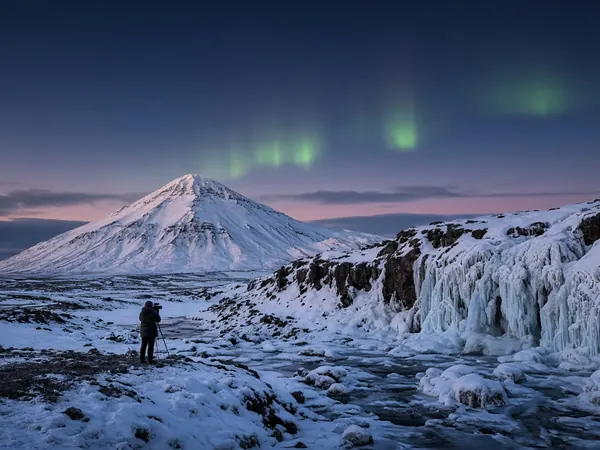 A cone-shaped snowy mountain and a frozen waterfall under faint northern lights, with a photographer